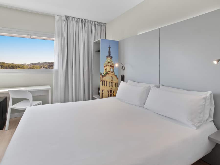 Quarto de hotel com uma cama grande, uma mesa e vista para um edifício através de uma janela.
