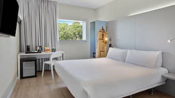 A modern hotel room with a large bed, a desk with a chair, a small refrigerator, and a window with curtains.
