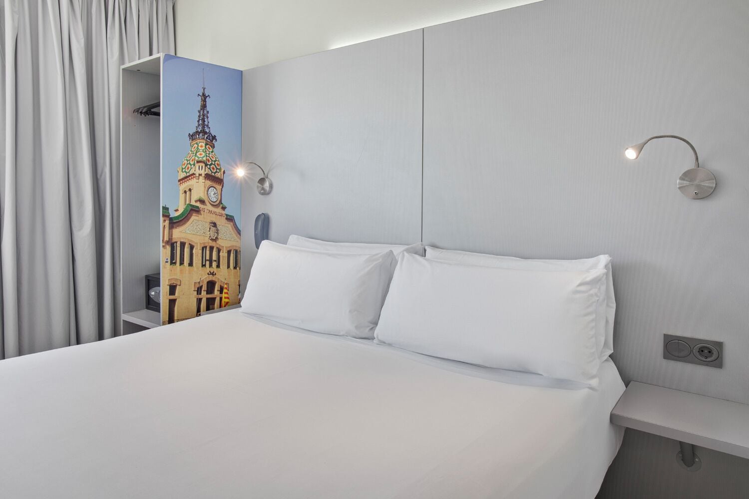 A hotel room with a large bed, two wall-mounted lights, a nightstand, and a wall with a colorful mural of a building.
