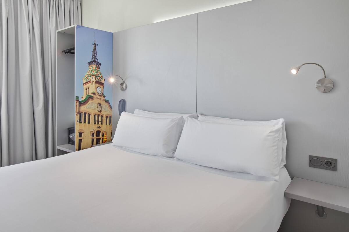 A hotel room with a large bed, white bedding, a wall-mounted light fixture, and a decorative panel with a building image.