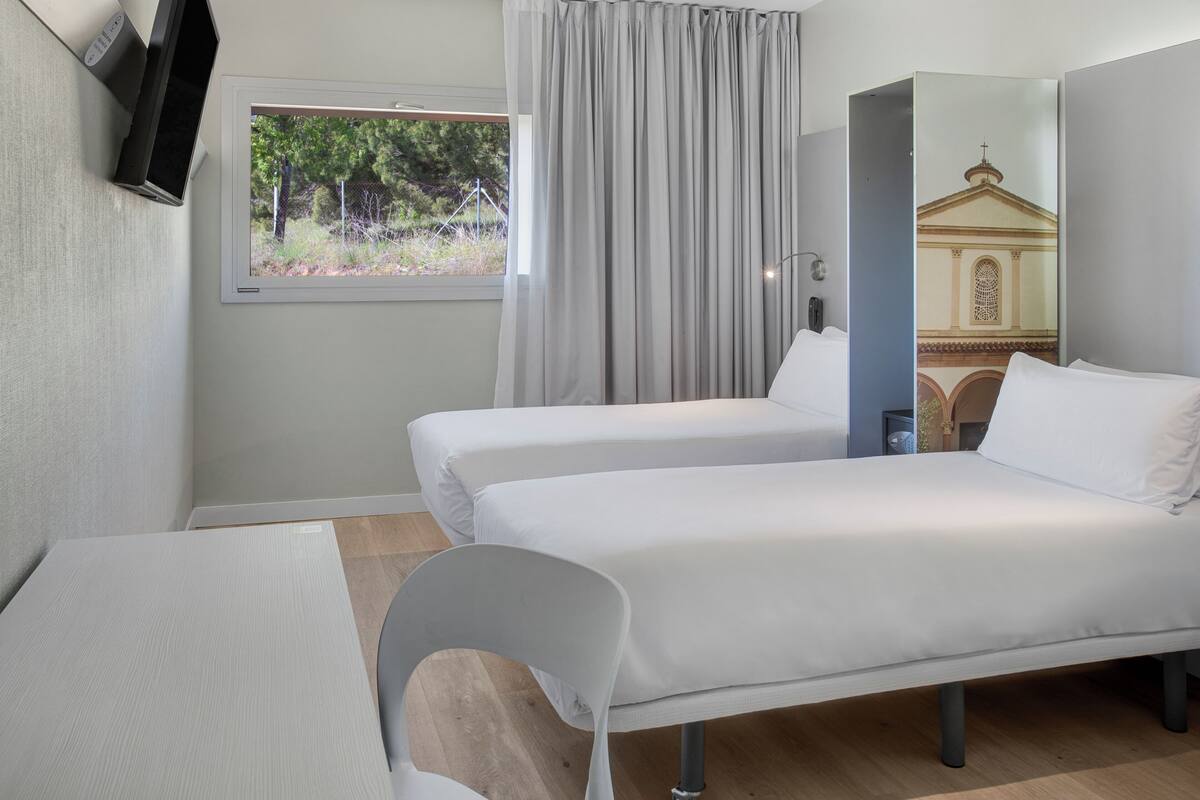 A hotel room with two beds, a desk, a chair, a TV, a window with curtains, and a mirror reflecting a building.