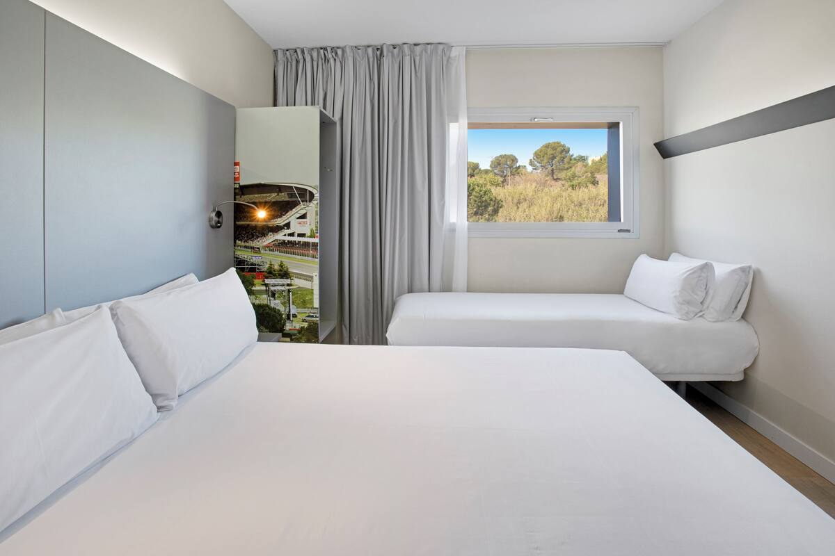 A hotel room with a large bed, a smaller bed, and a window with a view of trees and a stadium.