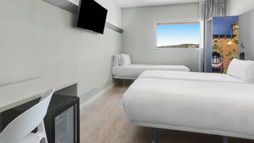 Habitación de hotel con dos camas, un televisor montado en la pared, un ventanal con vistas a la ciudad y un escritorio con una silla.