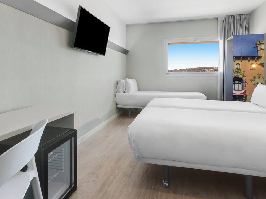 Habitación de hotel con dos camas, un televisor montado en la pared, un ventanal con vistas a la ciudad y un escritorio con una silla.
