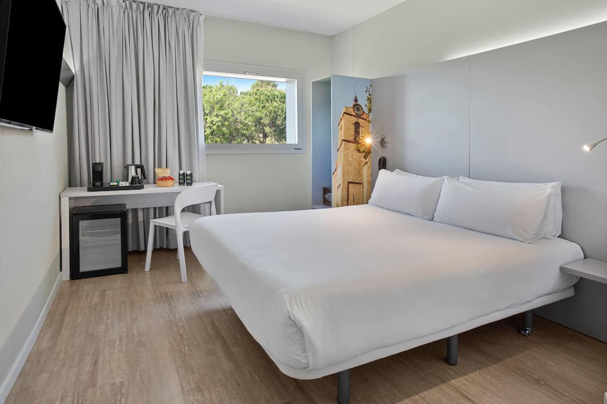 Habitación de hotel moderna con una cama grande, un escritorio con silla, un refrigerador pequeño y un televisor montado en la pared.