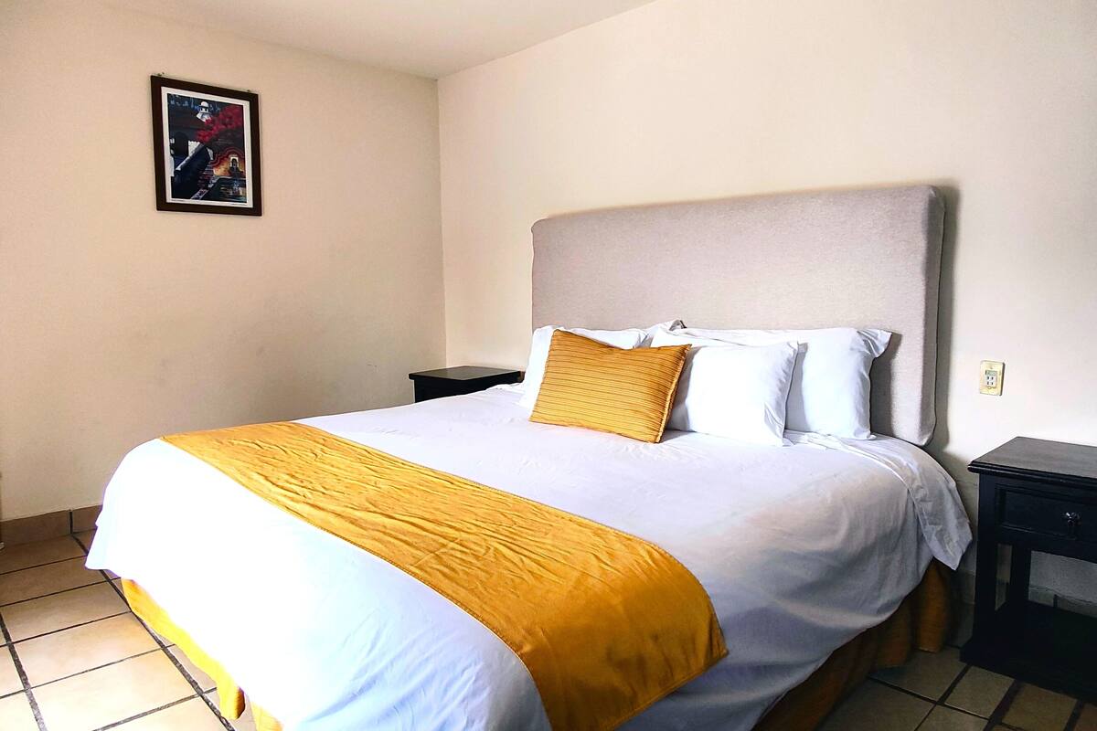 Ein ordentlich bezogenes Bett mit einer weißen und gelben Tagesdecke, einem einzelnen goldfarbenen Kissen und einem Kopfteil. Am Bett befindet sich ein kleiner, schwarzer Nachttisch mit einer Schublade.