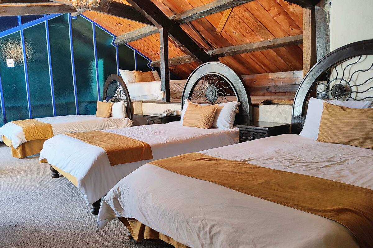 Ein Zimmer mit drei Betten, jeweils mit Holz Kopfteil und weißer Bettwäsche. Das Zimmer hat eine schräge Decke mit Holzbalken und einen Blick ins Freie durch große Fenster.