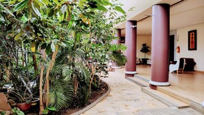 Un couloir au sol carrelé, bordé de plantes en pot et d’arbres, qui mène à un espace détente où l’on trouve un extincteur accroché au mur.