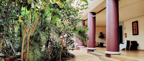 A spacious indoor area with a tiled floor, surrounded by lush green plants and tall red columns.