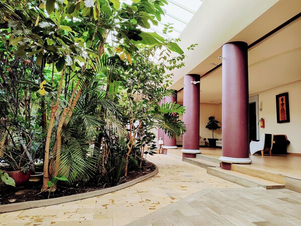 A spacious indoor area with a tiled floor, surrounded by lush green plants and tall red columns.
