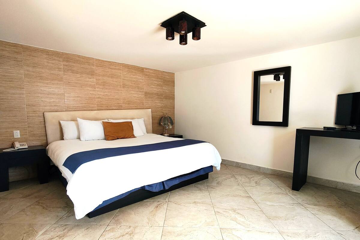 Ein Hotelzimmer mit einem großen Bett, einem Nachttisch mit Telefon, einem Schreibtisch mit Fernseher und einem Spiegel an der Wand.