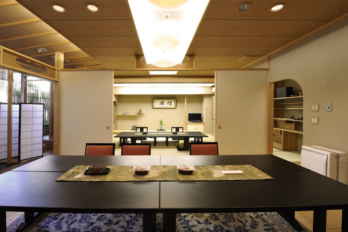 A dining area with a long table, chairs, and a view of a kitchen in the background.