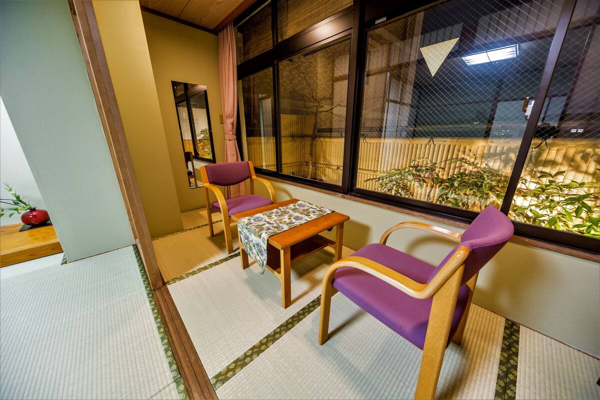 A traditional Japanese room with tatami flooring, a low wooden table, purple and wooden chairs, and a view of bamboo outside.