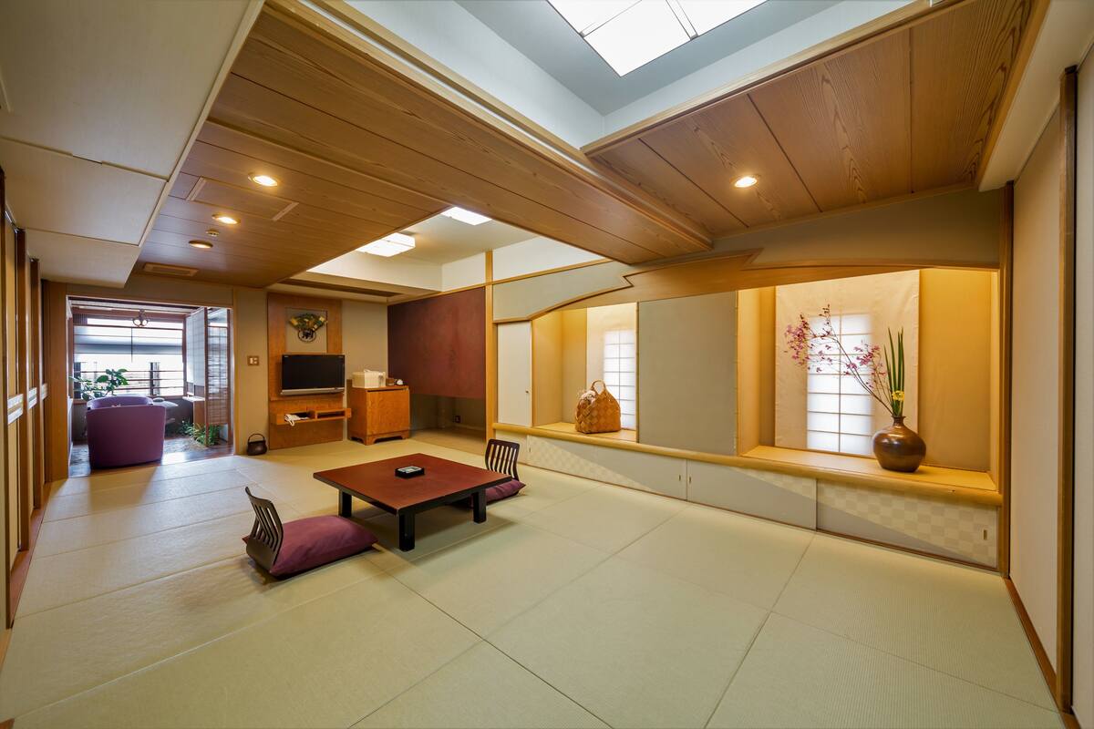 A traditional Japanese-style room with tatami flooring, a low table with cushions, a TV, and a window with a view of the outside.