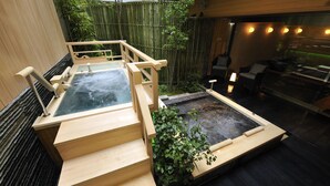 Zona de baños termales con escalones de madera que conducen al agua, rodeada por una valla de bambú y un pequeño jardín.