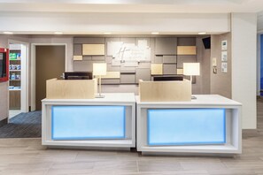 Un comptoir d’accueil au design moderne, doté de deux guichets avec des panneaux bleus rétroéclairés, d’une enseigne fixée au mur au-dessus, et d’un petit réfrigérateur placé à gauche.