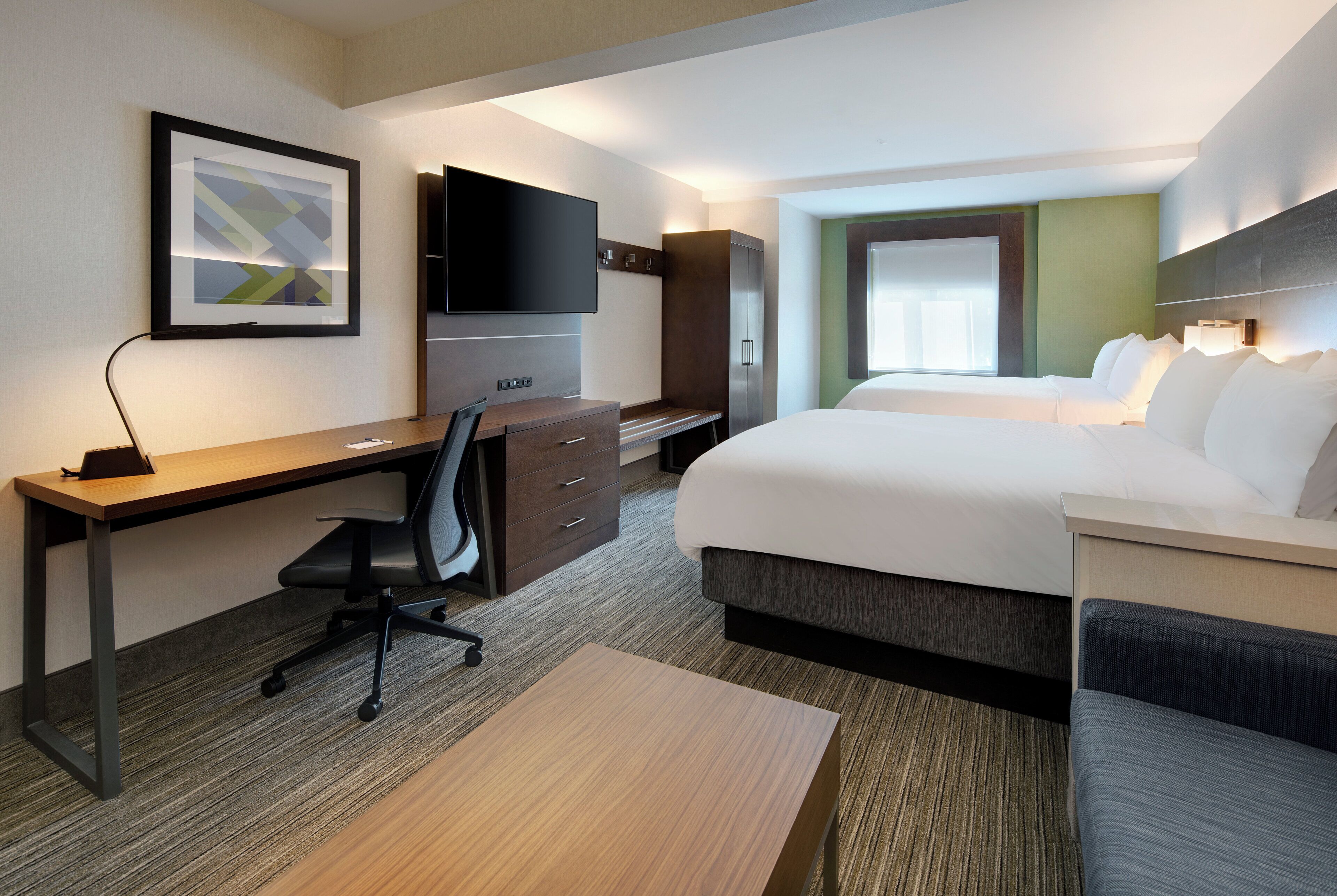 Quarto de hotel com cama, escrivaninha, cadeira, televisão e um quadro na parede.