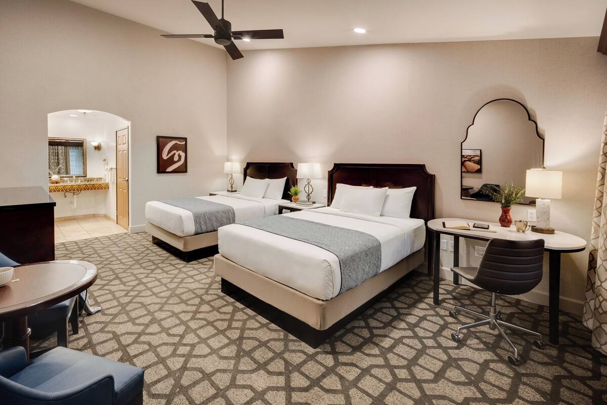 Habitación de hotel con dos camas, un escritorio, una silla, ventilador de techo y baño visible a través de una puerta abierta.