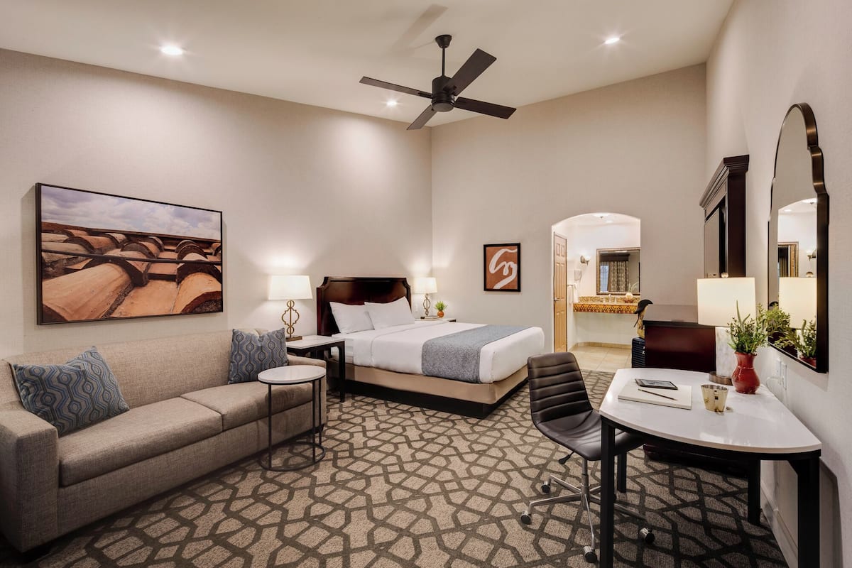 Habitación de hotel con cama, sofá, escritorio, silla, ventilador de techo y un cuadro grande en la pared.