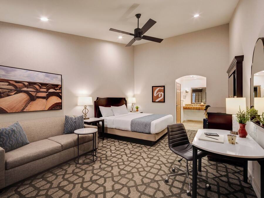 A hotel room with a bed, a sofa, a desk, a chair, a ceiling fan, and a large painting on the wall.