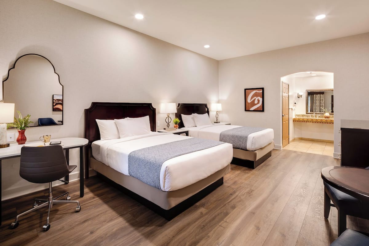 Habitación de hotel con dos camas, un escritorio, una silla, una mesita y un baño visible a través de una puerta abierta.