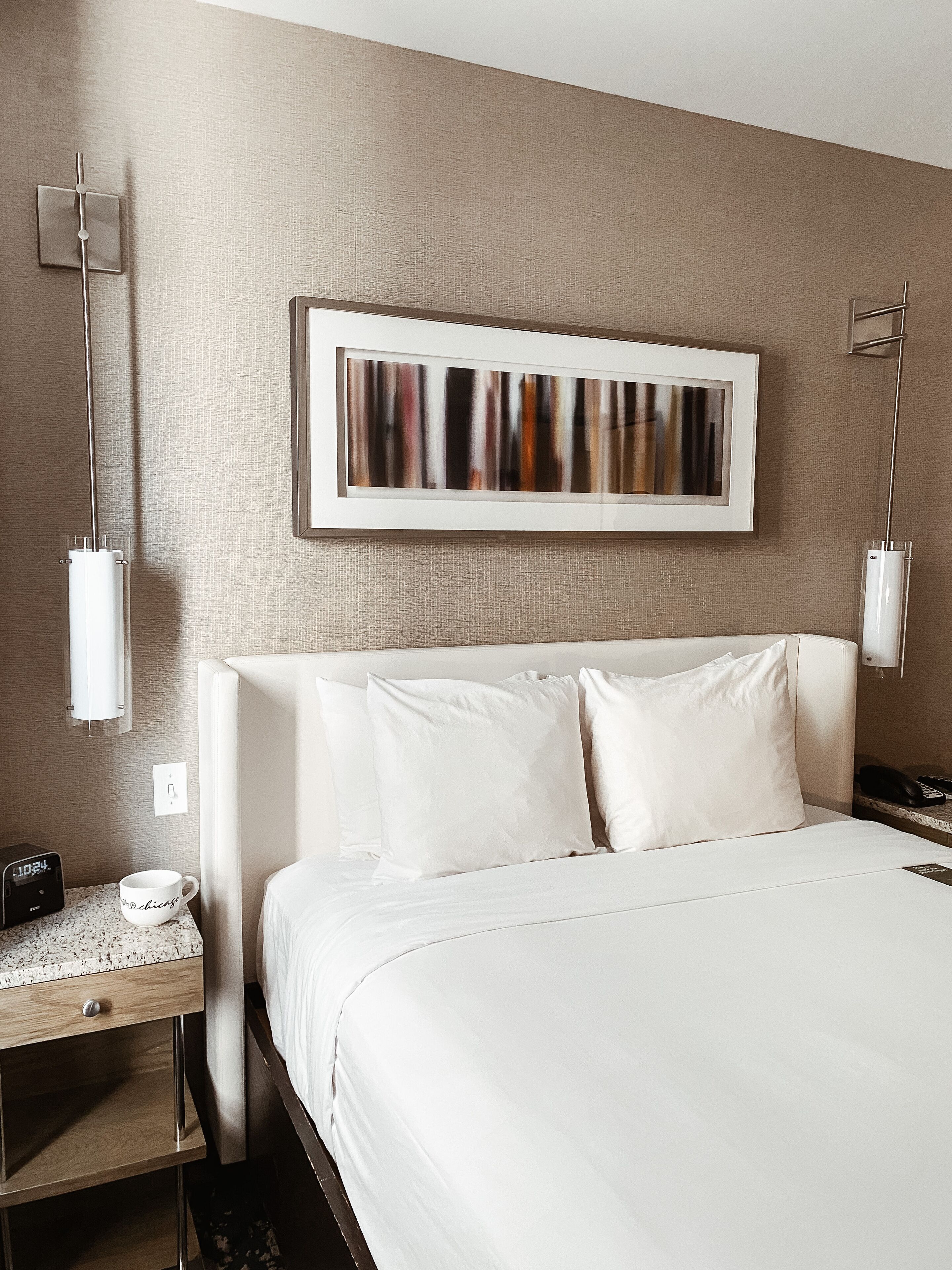 Habitación de hotel con una cama grande, una mesita de noche con un teléfono y una taza, y un cuadro abstracto colgado en la pared.