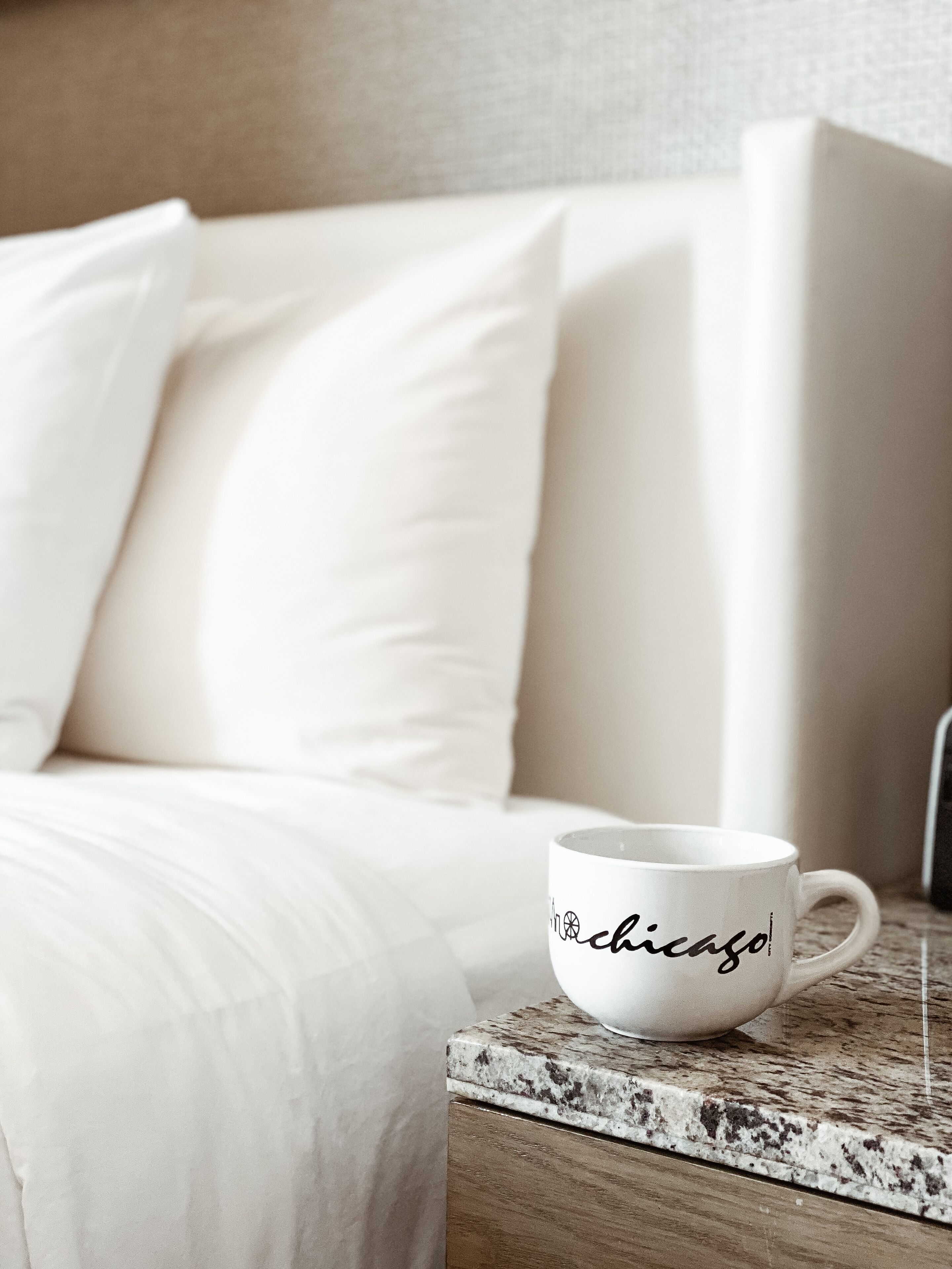 A white mug with "Chicago" written on it, placed on a wooden side table next to a bed with white bedding.