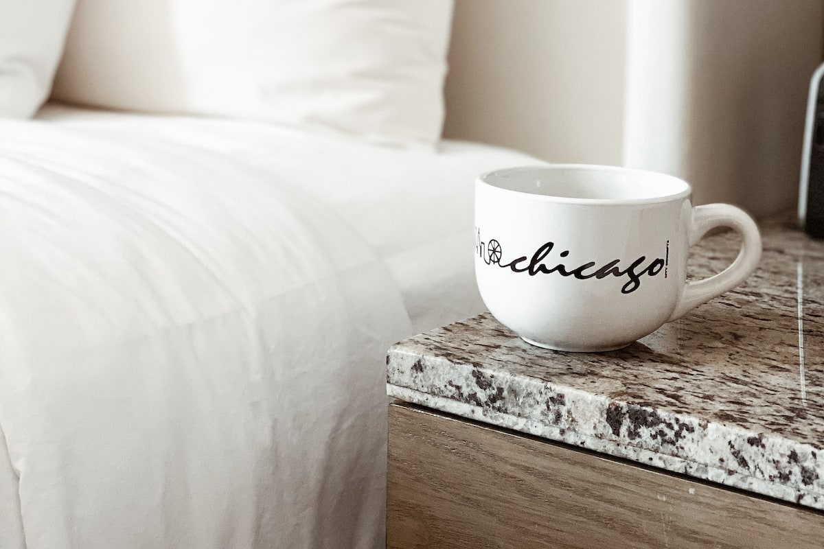 A white mug with "Chicago" written on it, placed on a wooden side table next to a bed with white bedding.