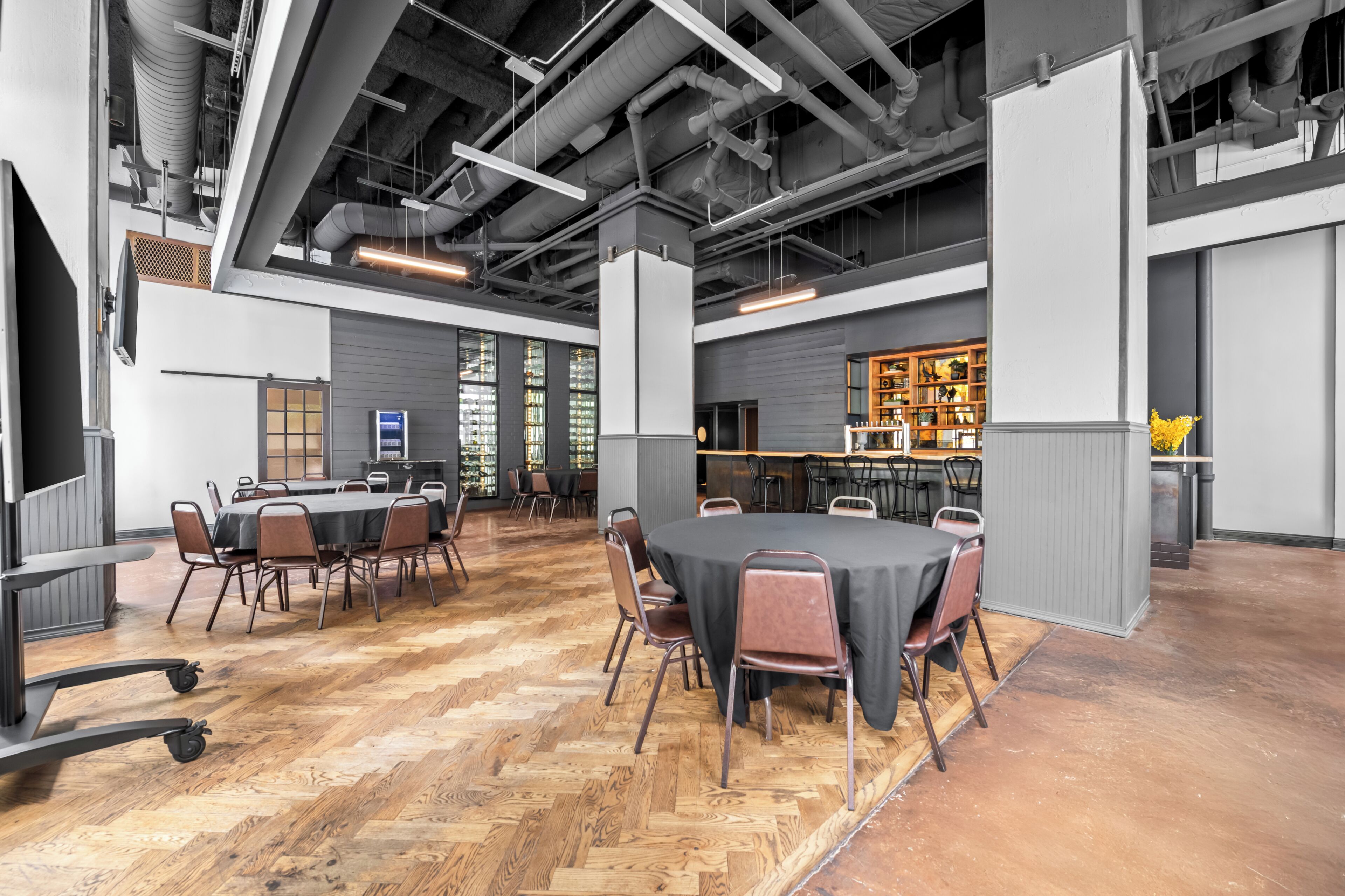A spacious conference room with a large table and chairs, a bar area with shelves, and a herringbone-patterned wooden floor.
