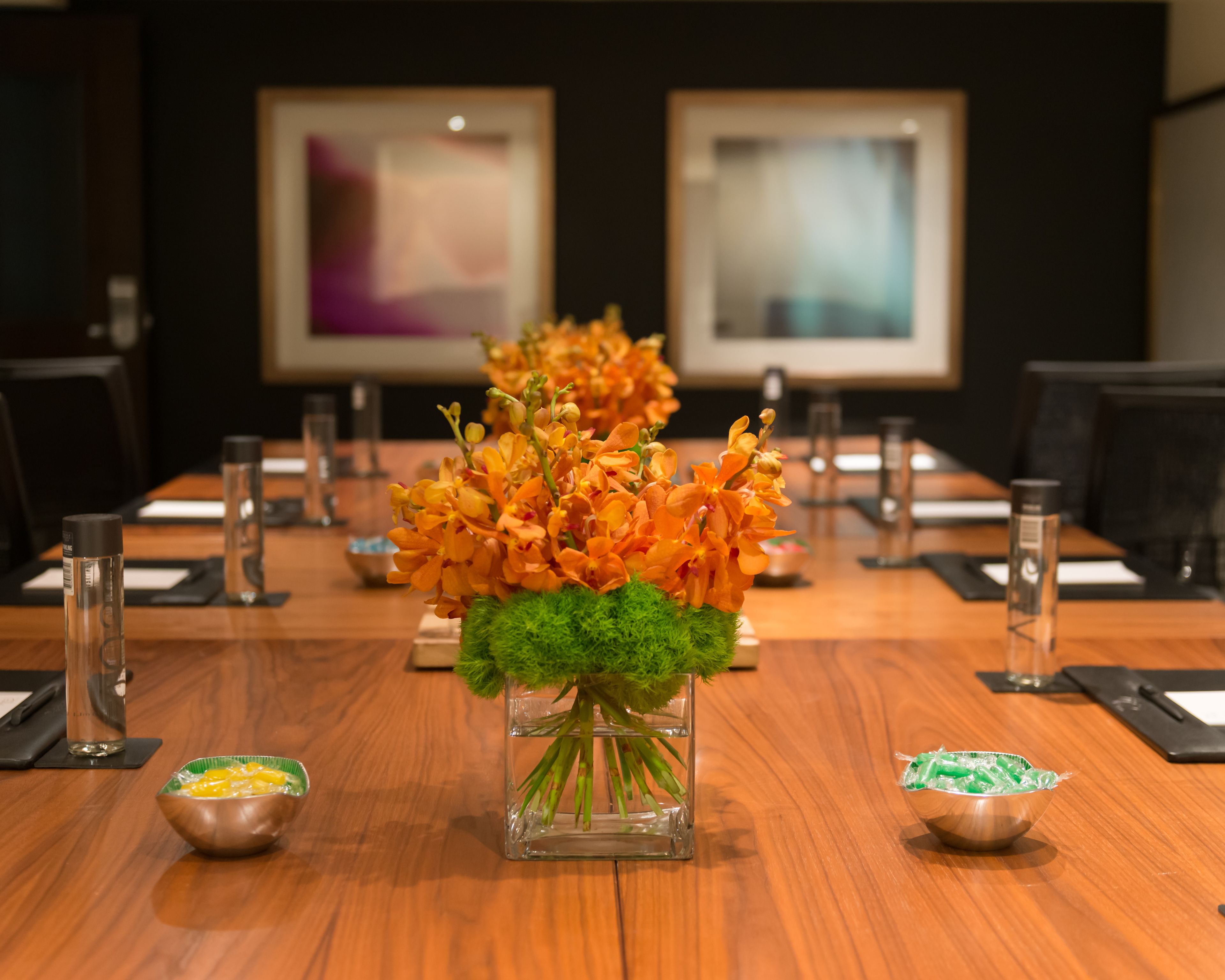 Una mesa de conferencias con un centro de mesa floral, botellas de agua y pequeños cuencos sobre una superficie de madera.