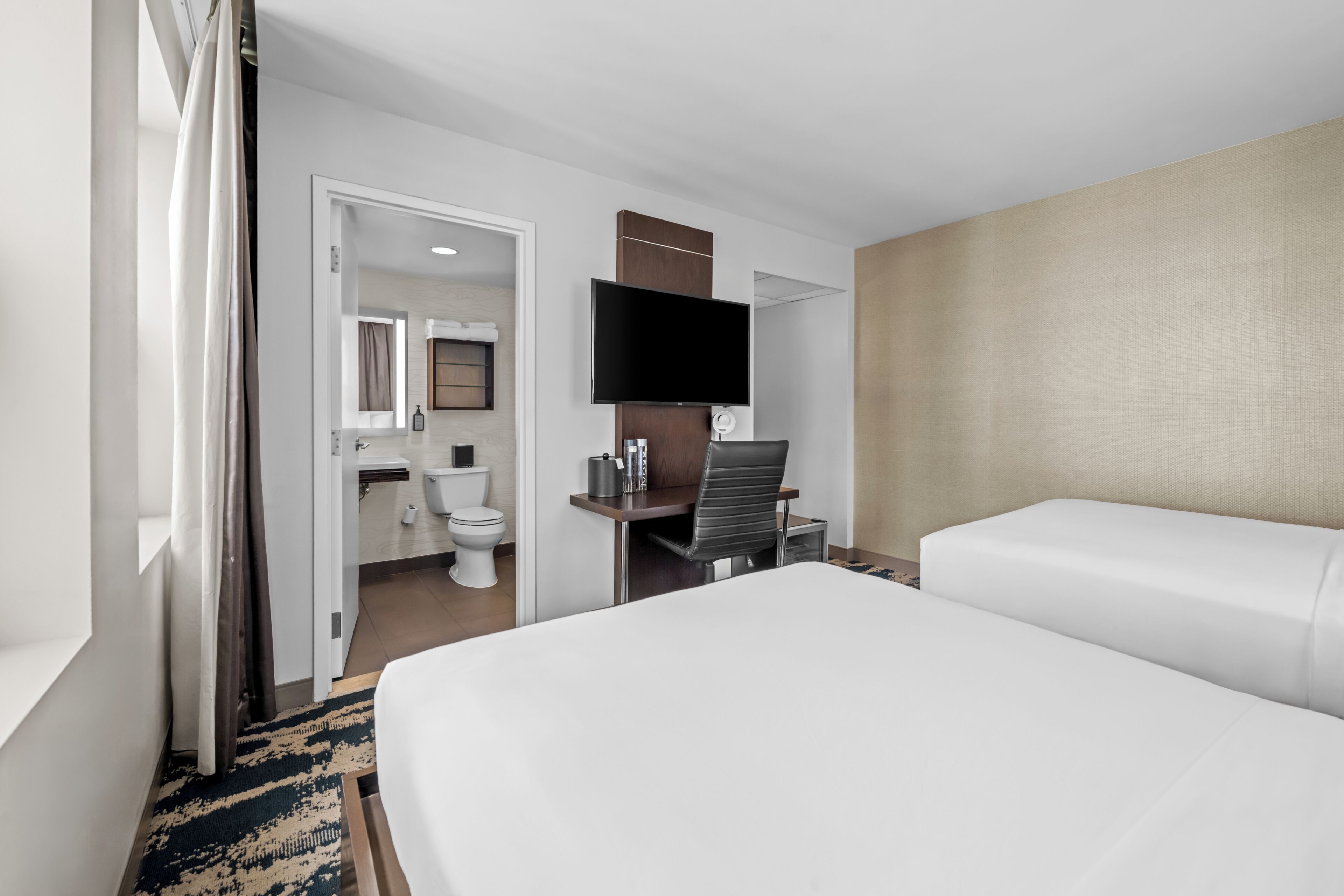 A hotel room with a bed, a television, a desk, and a bathroom visible through an open door.