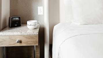 Une chambre d’hôtel avec un lit, une table de chevet avec une horloge et une tasse, une lampe fixée au mur et un tableau au-dessus du lit.