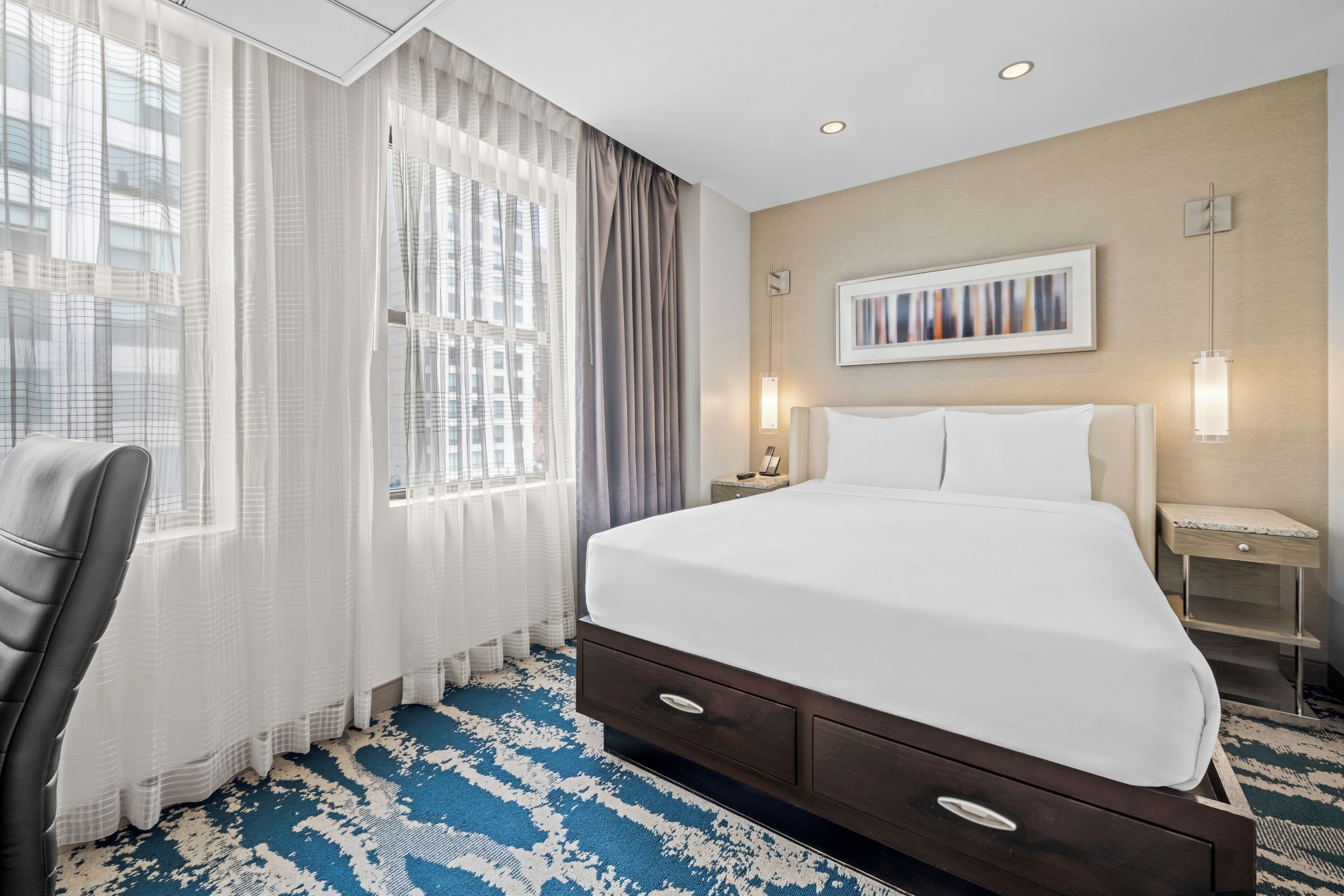 A hotel room with a bed, a chair, a nightstand, a lamp, and a painting on the wall.