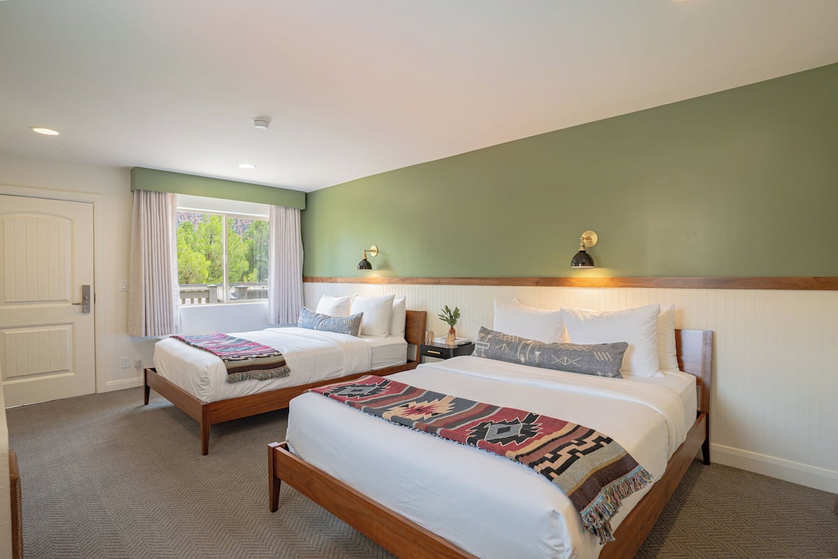 A hotel room with two beds, a large window with curtains, a nightstand with a lamp, and a view of greenery outside.