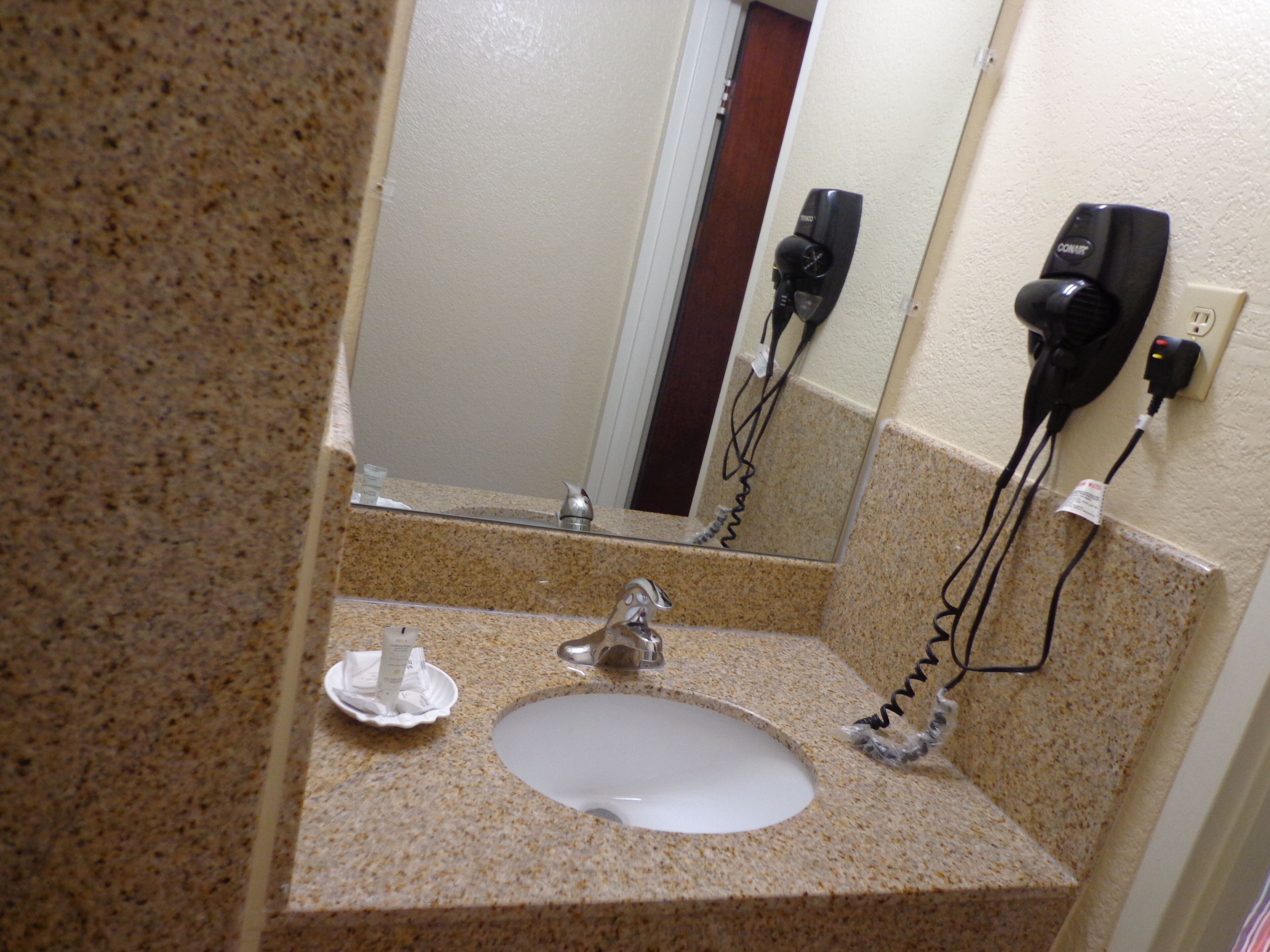 A bathroom with a granite countertop, a sink, a mirror, and a hairdryer mounted on the wall.
