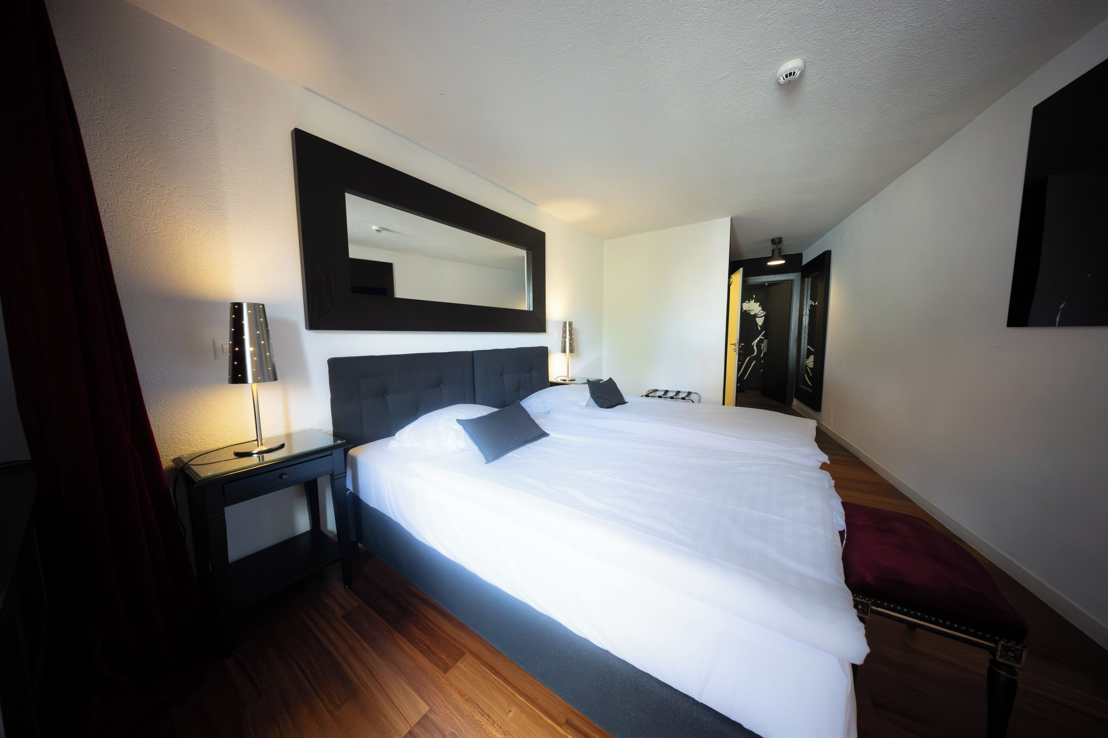 A modern hotel room with a large bed, a nightstand, a mirror, and a red bench.
