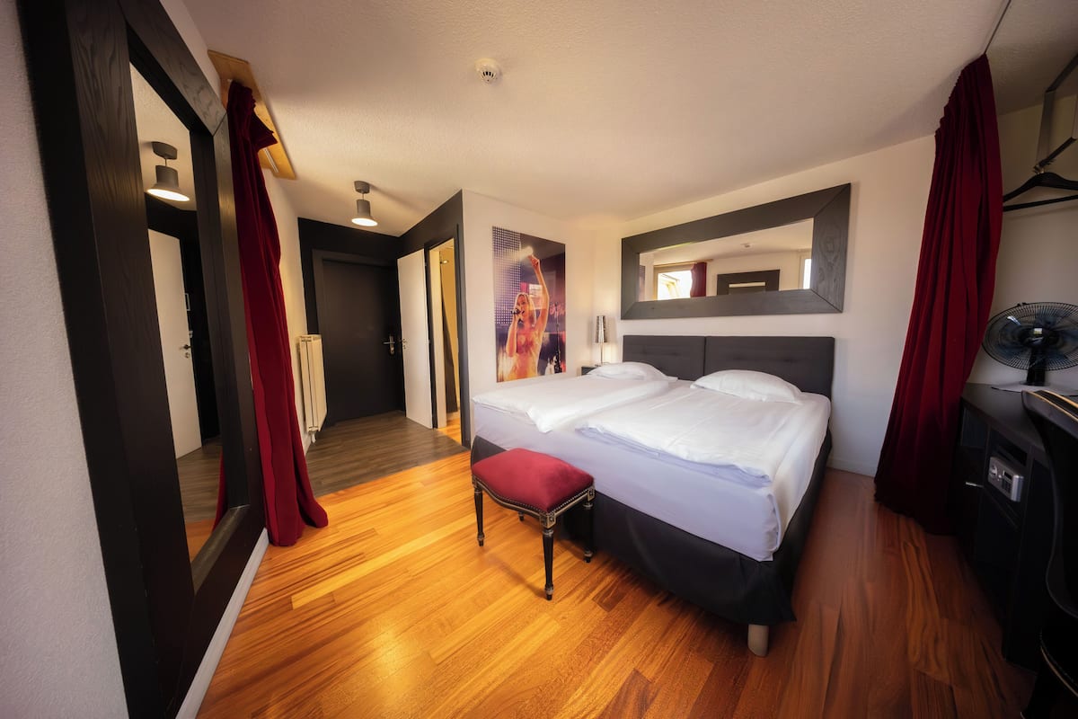 A hotel room with a double bed, a small red bench, a bedside table, a mirror, and a fan.