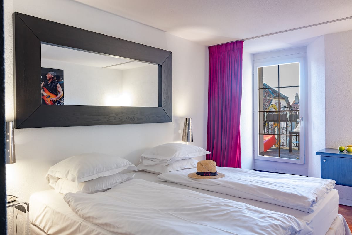 A hotel room with a large mirror, a bed with white linens, a hat on the bed, a window with pink curtains, and a view of buildings outside.
