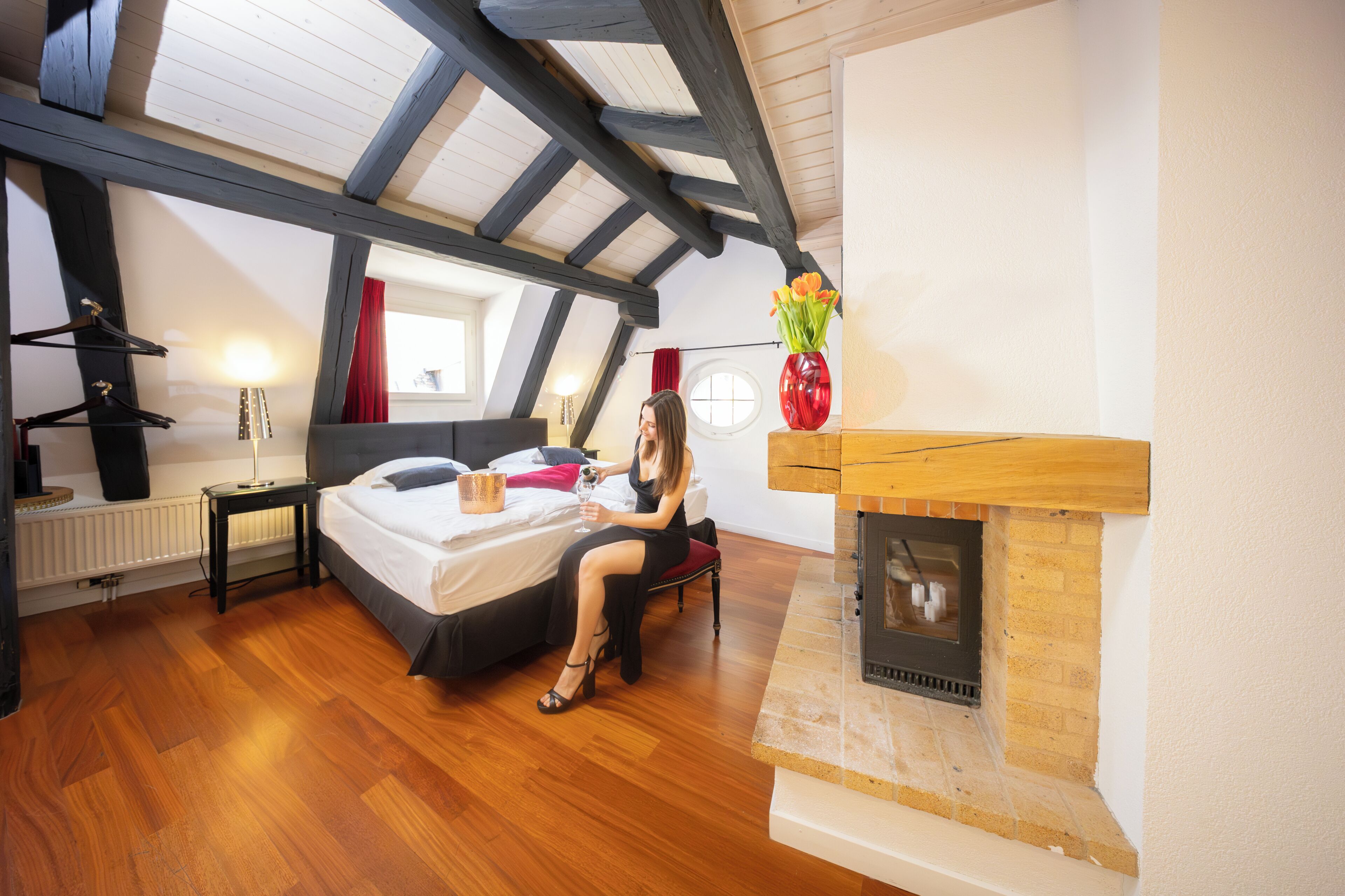 A bedroom with a bed, a bedside table, a red vase with flowers, a fireplace, and a skylight.