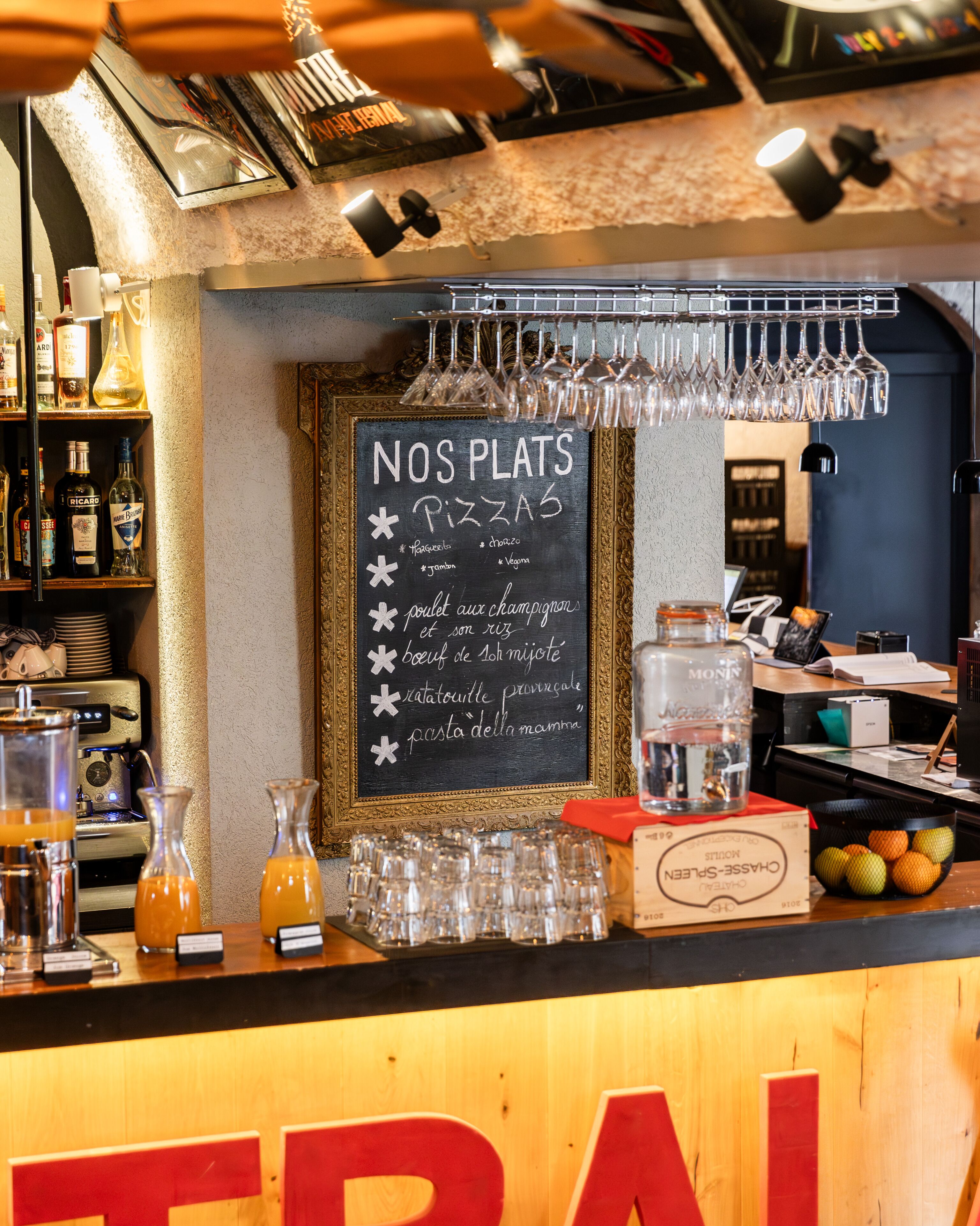 A bar area with a chalkboard menu listing pizza options, glasses, bottles, and a cash register.