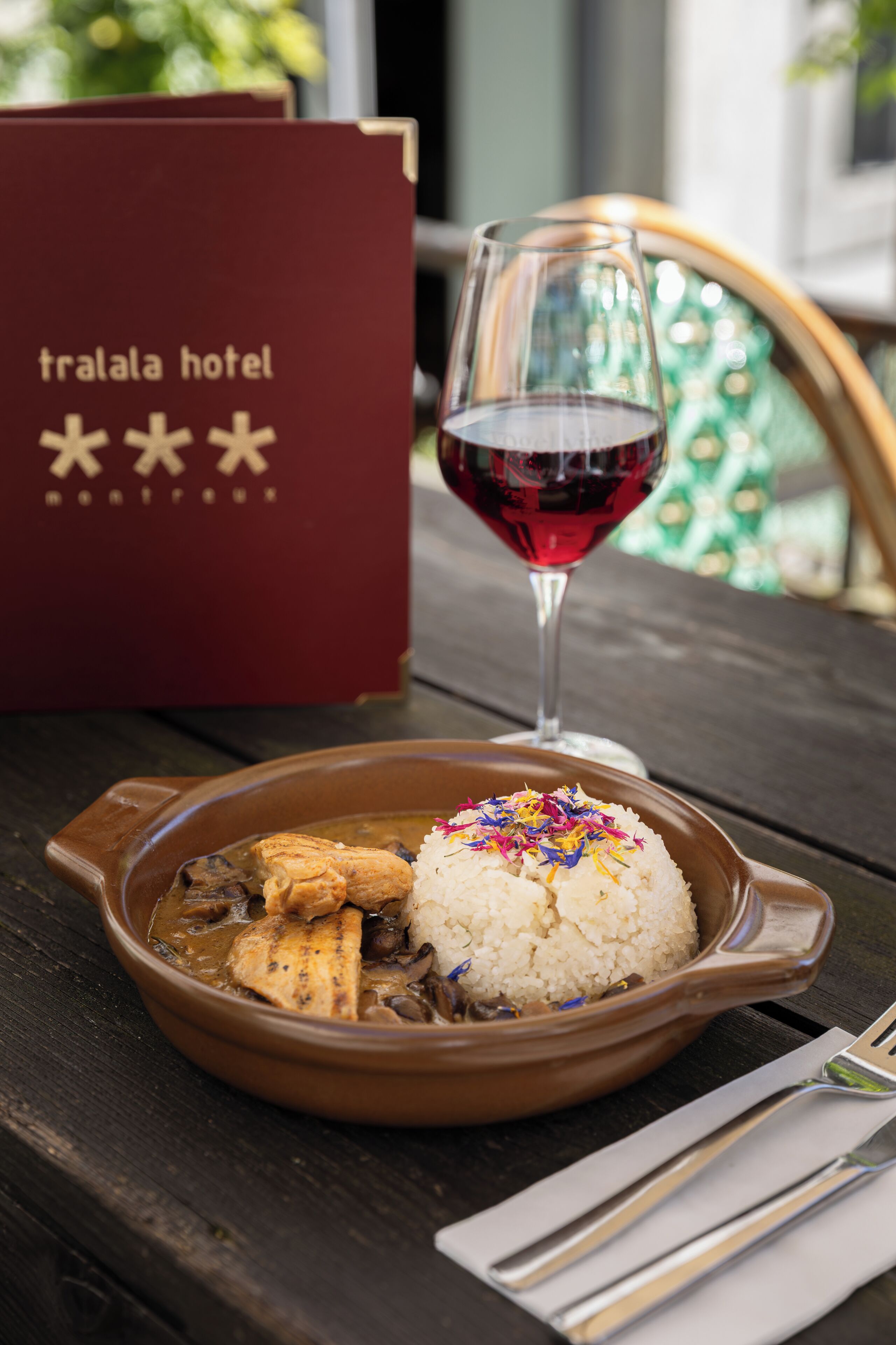 A table with a brown dish containing rice and chicken, a glass of red wine, and a red hotel brochure.