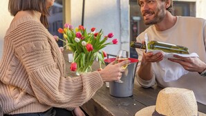 Two people seated at an outdoor table with a bottle of wine, a glass, and a vase of tulips.