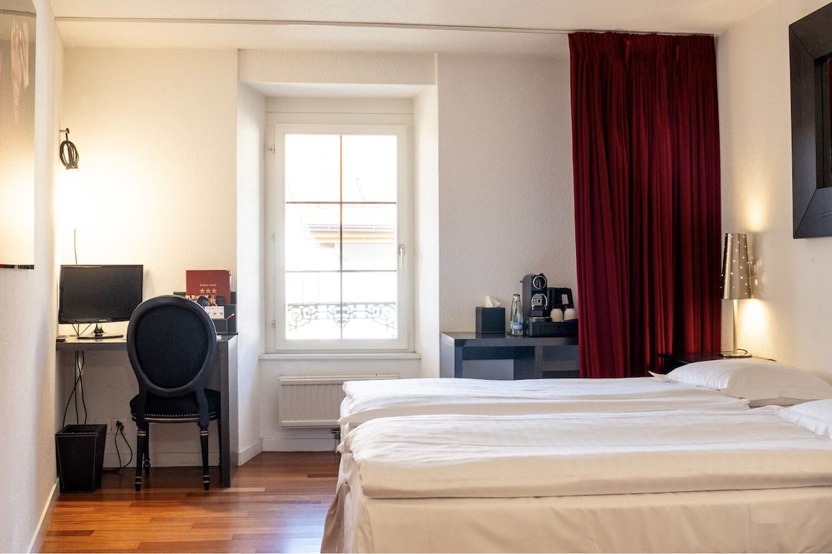A hotel room with a bed, a desk with a computer, a chair, a window with curtains, and a bedside table with a lamp.
