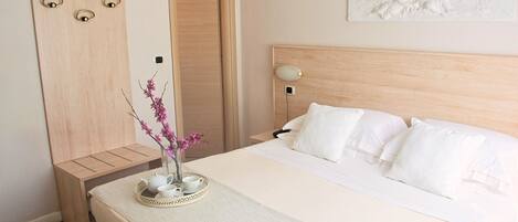 A neatly made bed with white bedding, a bedside table with a vase of pink flowers, and a wall-mounted artwork.