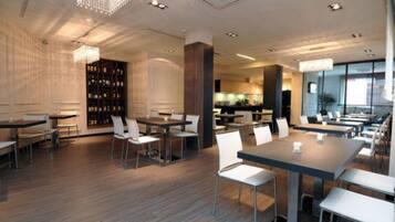 A modern dining area with wooden flooring, white chairs, and tables. There is a bar area with a wine rack in the background.