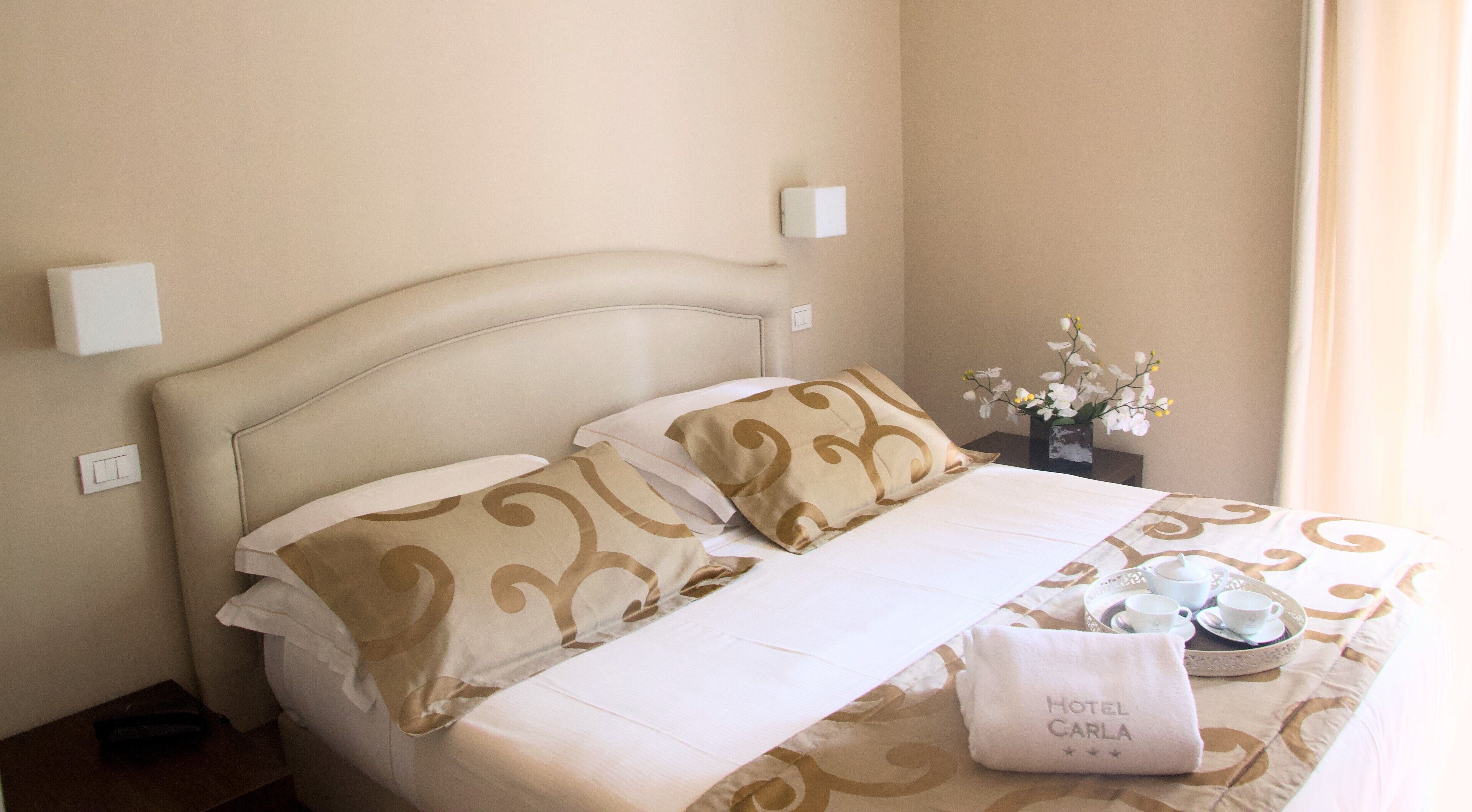 A hotel room with a bed, pillows, a nightstand with a vase of flowers, and a tray with cups and a teapot.