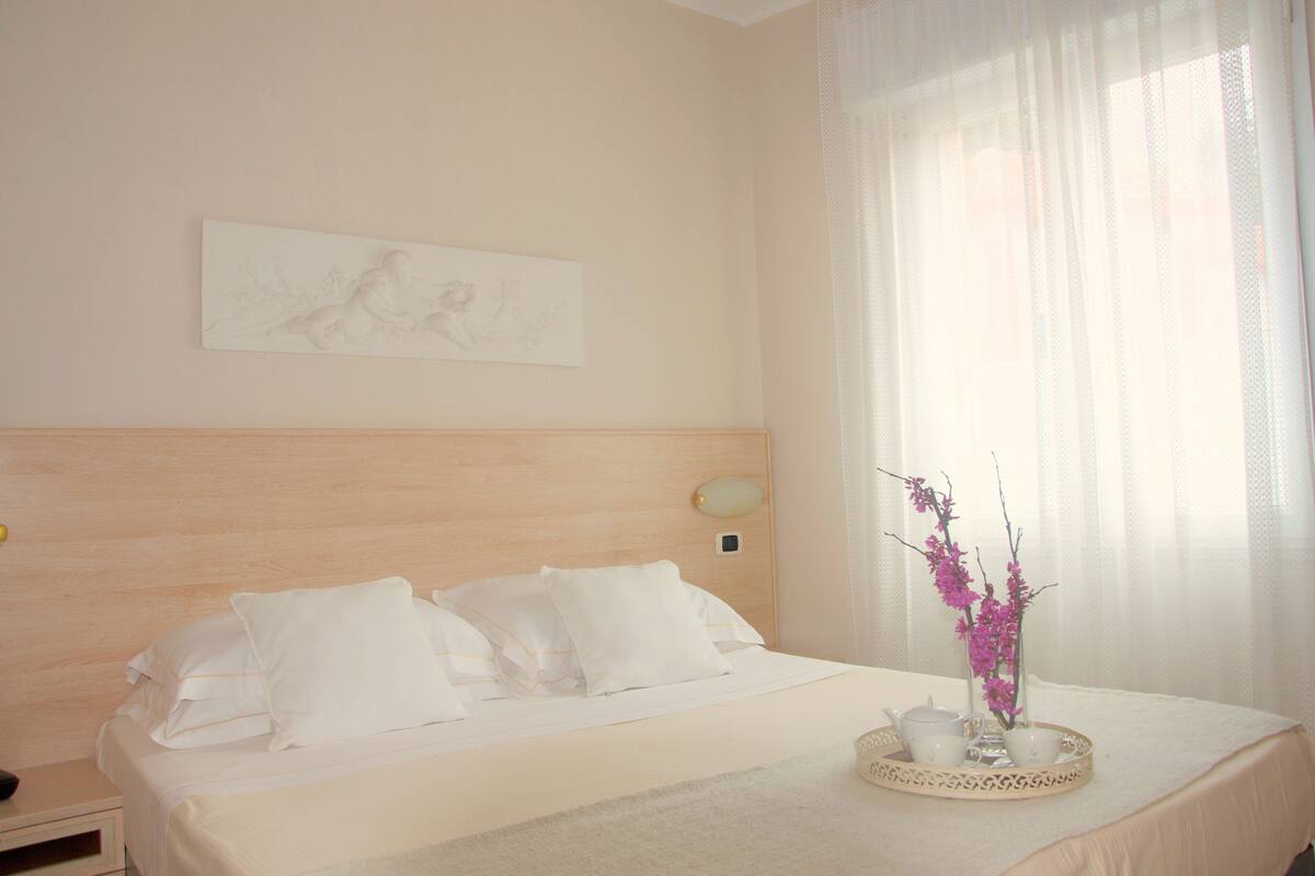 Una cama bien hecha con sábanas y almohadas blancas, una mesita de noche con lámpara, una obra de arte en la pared y una ventana con cortinas de encaje.