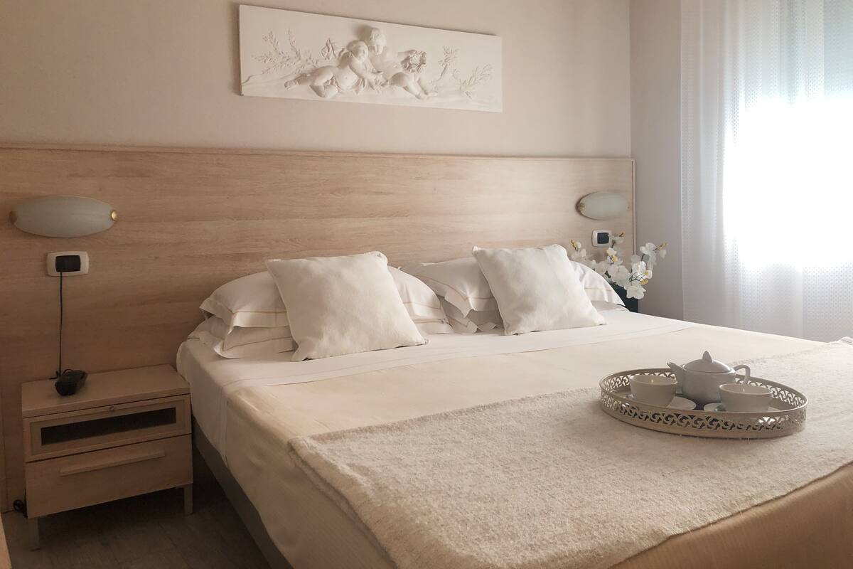 Una cama bien hecha con ropa blanca, una mesita de noche con una lámpara y una bandeja con juego de té.