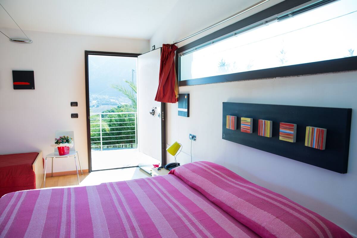 A bedroom with a bed, a chair, a small table, a window, and a view of greenery.