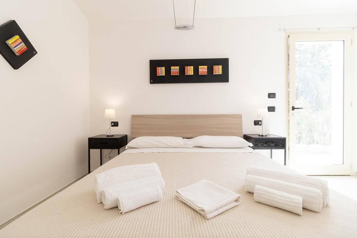 A modern hotel room with a large bed, two bedside tables with lamps, a wooden headboard, and a view of greenery through the door.